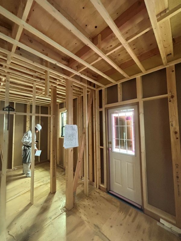 Interior of a house under construction with exposed wooden studs, ceiling joists, and a worker in a hard hat near a doorway.