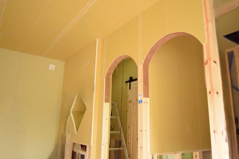 Interior of a building under construction with yellow drywall, arched doorway frames, and a ladder nearby.