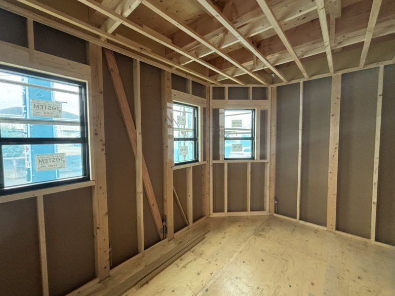 Interior of a room under construction with exposed wooden studs and framing, several windows, and unfinished floorboards.