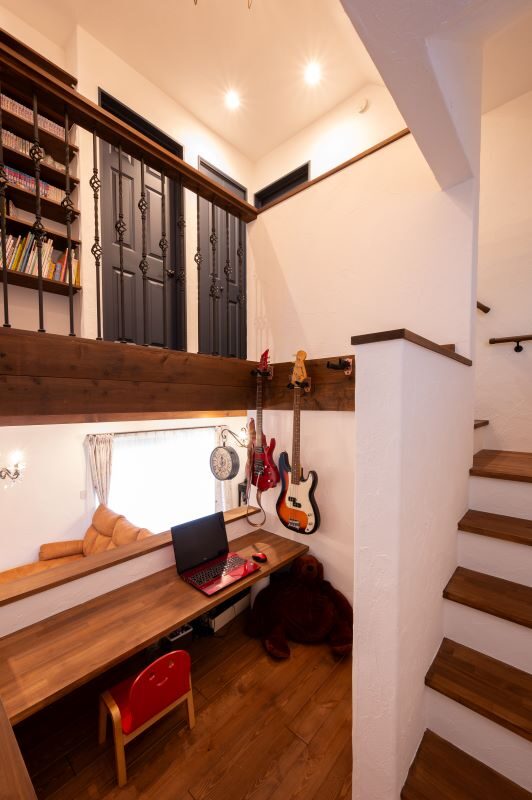 Cozy home workspace under a loft: wooden desk with laptop, red chair, guitar wall decor, and a staircase to the right.