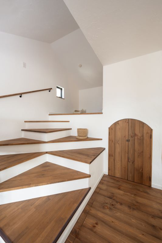 Wooden staggered staircase with white risers curves up along a white wall, small arch-shaped wooden door on the right, warm wood tones throughout.