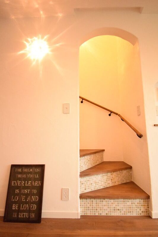 Cozy entryway with an arched doorway opening to a tiled staircase with a wooden handrail and warm overhead light; a leaning sign shows a love quote.
