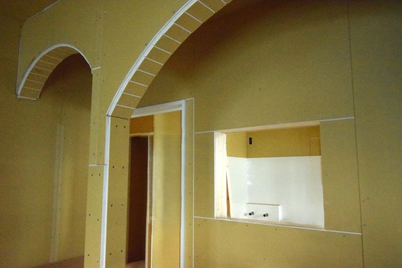 Interior under construction: yellow drywall with arched doorway and a rectangular pass-through to another room, visible framing and unfinished walls.