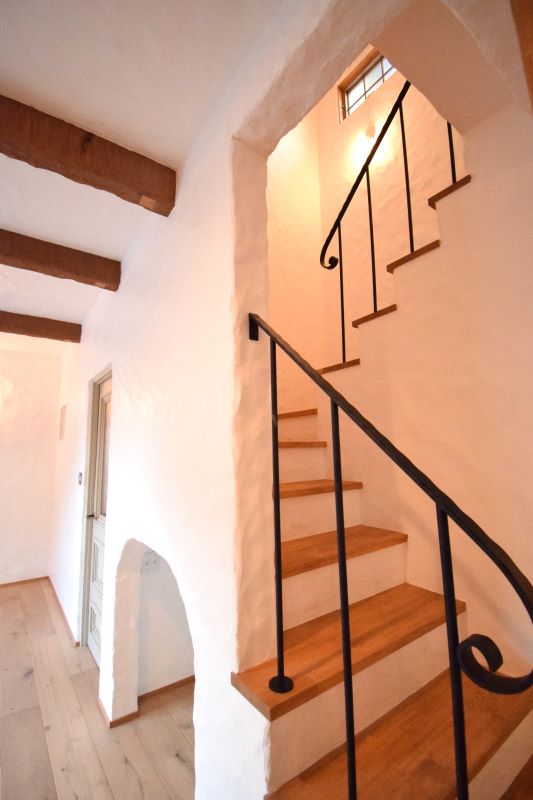 Interior staircase with wooden steps and black metal balusters winding up beside white plaster walls and exposed wooden ceiling beams, in a sunlit hallway.