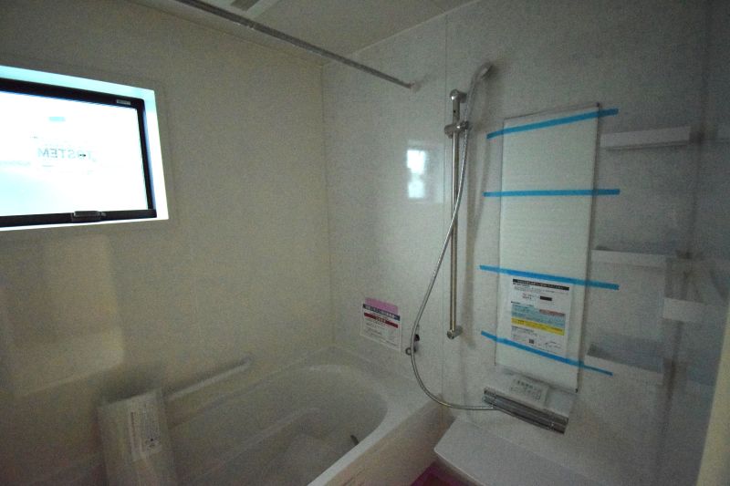 White bathroom featuring a built-in bathtub with a handheld shower; window on the left and blue painter's tape protecting a glass panel on the right edge of the room.
