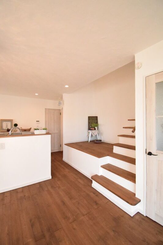 Bright open living area with white walls, light fixtures, a kitchen island on the left, and a raised wooden-step staircase on the right leading to a platform.