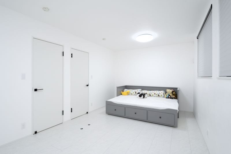 White minimalist bedroom featuring a gray bed with storage drawers at the foot, two doors on the left, and windows with blinds on the right side.