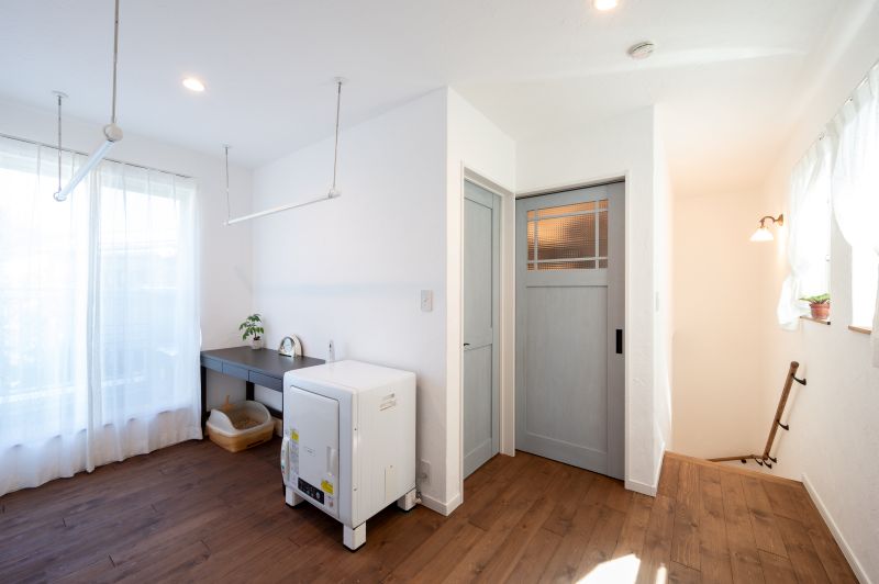 Bright laundry nook with a top-loading washing machine on a wooden floor, gray counter, small plant, and sheer-curtained window letting in light.