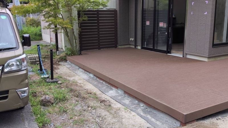 Brown raised deck extends from the house to sliding glass doors, with a dark privacy fence at the far end and surrounding plants and dirt nearby.