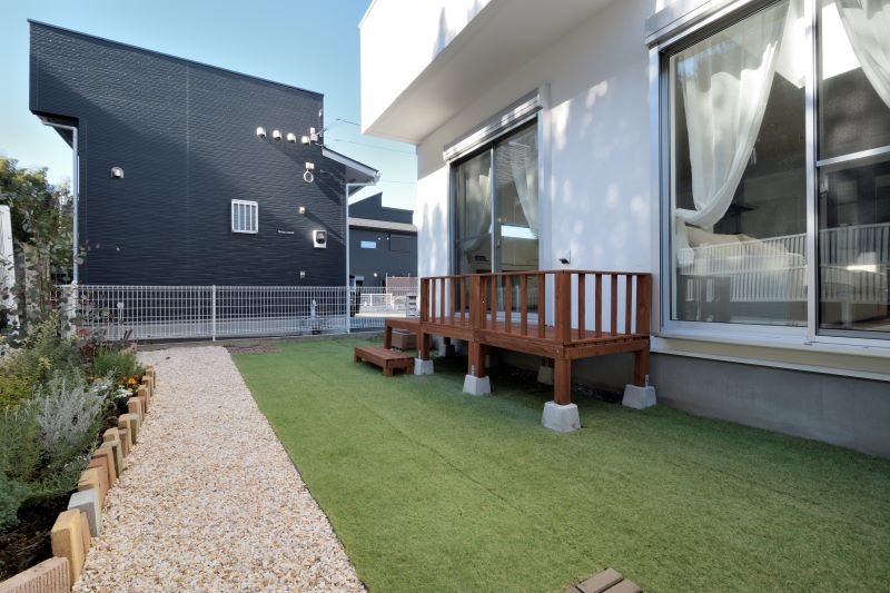 Backyard of a white modern house with a raised wooden deck on concrete supports, a gravel path, and artificial turf.