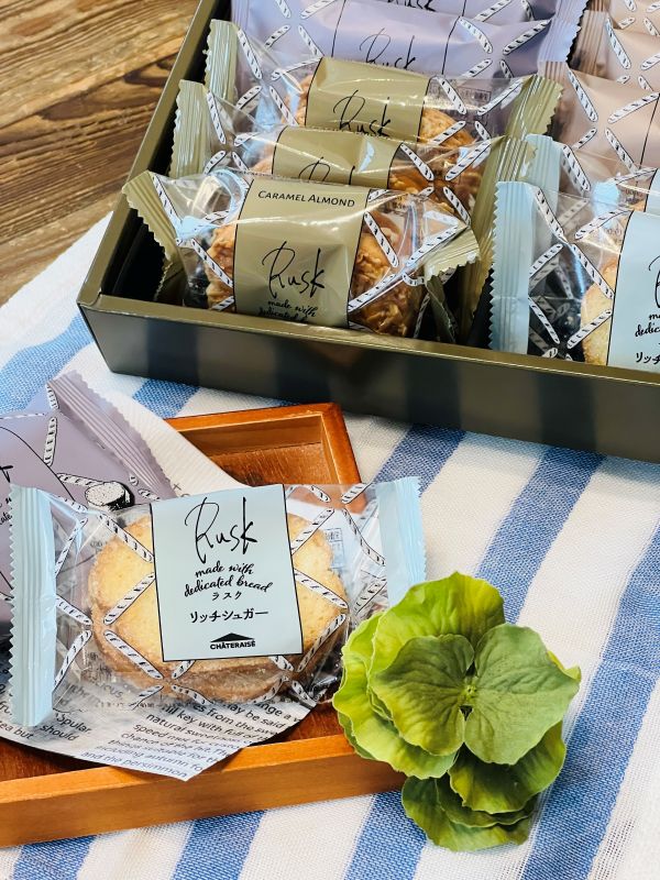 Gift box of individually wrapped rusk biscuits with caramel almond flavor; pastel wrappers and blue striped cloth backdrop on a wooden surface.