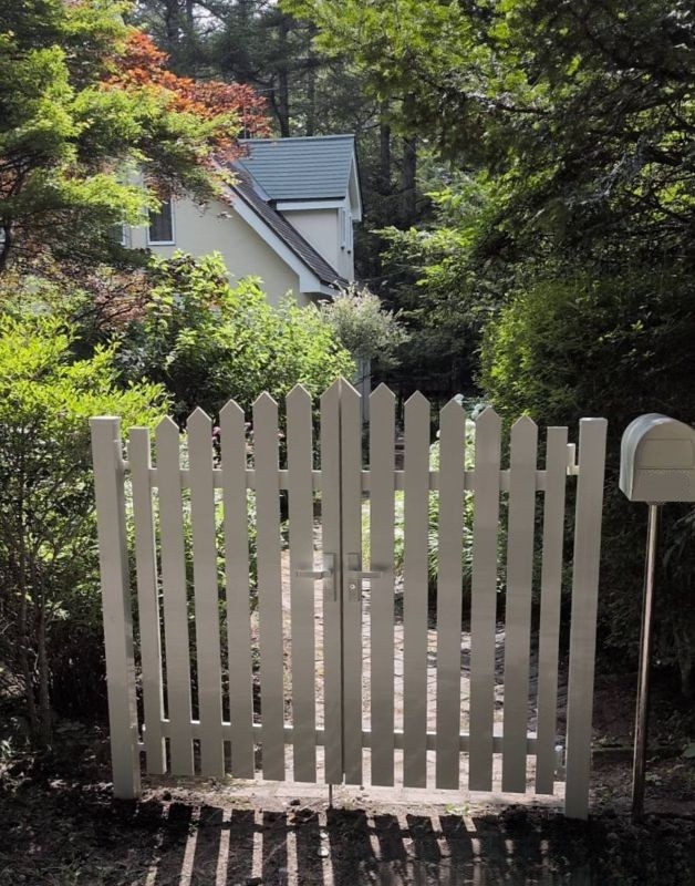 White wooden picket gate blocking a garden path toward a cream house among trees. (informative)