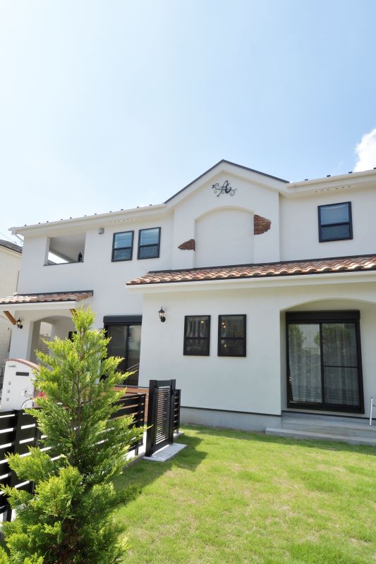 White two-story house with a red-tiled roof, black-framed windows, and a small fenced green yard under a clear blue sky