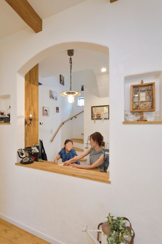 Mother and child sit at a wooden kitchen counter seen through a rounded archway in a white-walled home with stairs and wall photos in the background.