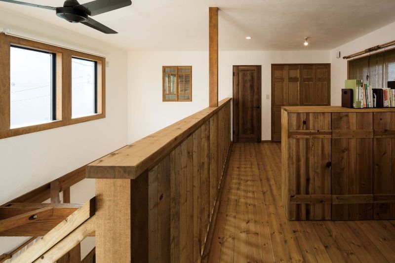 Rustic wooden loft hallway with a railing, exposed staircase, two windows on the left, and a wooden divider on the right.