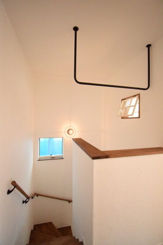 Interior stairwell with white walls, wooden steps and railing, a small square window, wall light, and a black U-shaped pipe overhead.