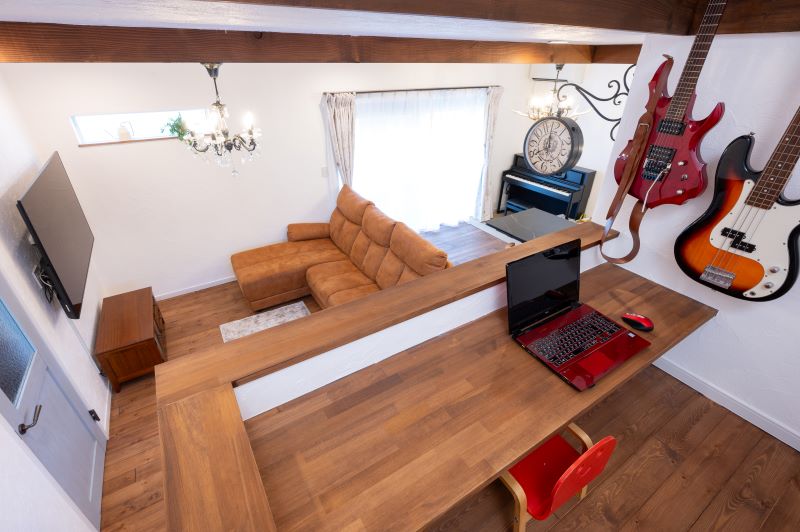 Loft living area with wooden beams, a brown sectional sofa, wall TV, chandelier, and guitars hanging on the white wall.