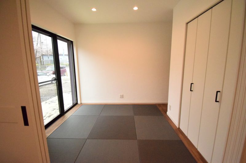 A small minimalist room with a tatami-style mat floor, large windows along one wall, and white built-in closets with black handles on the right.