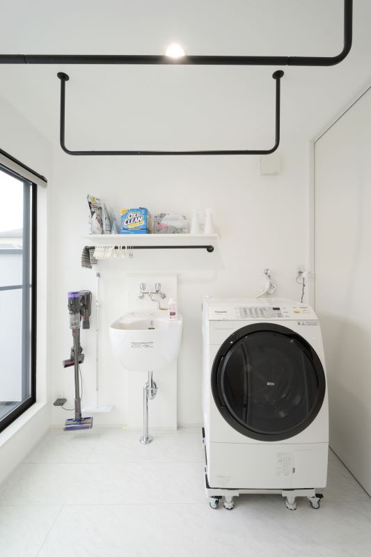 Bright laundry room with a front-loading washing machine beside a utility sink, shelves with detergent, and a vacuum on the floor.