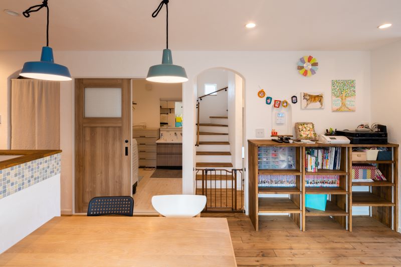 Bright playroom with a wooden table and white chair, blue/green pendant lights, open bookshelves, colorful wall art, and a gated staircase in the background.