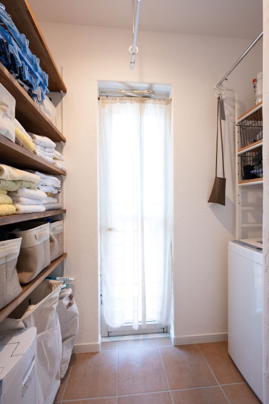 Narrow laundry room with tall shelves on the left holding towels and baskets, a washing machine on the right, and a sheer-curtained door at the back letting light in.