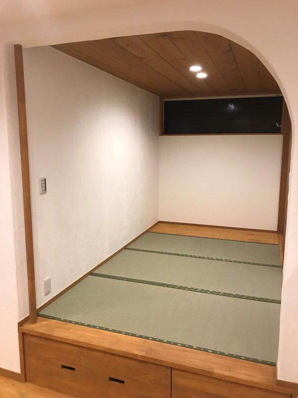 Raised tatami room with a wooden ceiling, arched doorway, and built-in storage drawers at the base; three tatami mats cover the floor and a small window sits high on the far wall.