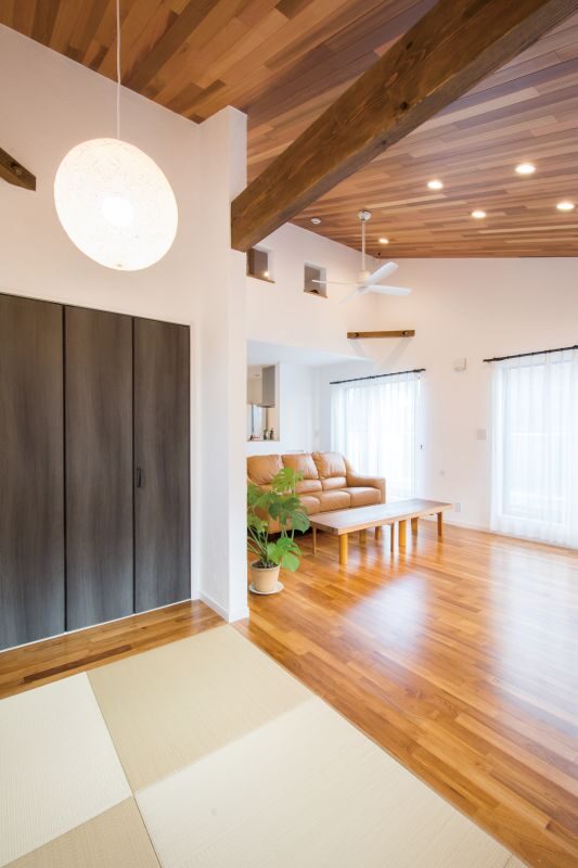 Bright modern living room with wooden ceiling beams, dark closet doors, tan leather sofa, low coffee table, potted plant, and large glass doors to the outside.