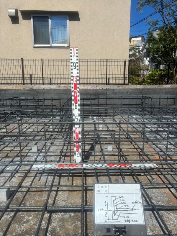 Construction site foundation with exposed steel rebar forming a grid; red-and-white measuring rods placed vertically and horizontally for alignment; a plan board sits nearby showing notes.