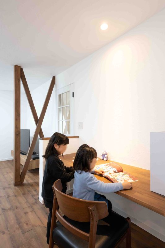 Two children sit at a wooden desk reading a magazine in a bright, minimalist room with a wooden X-shaped beam in the background