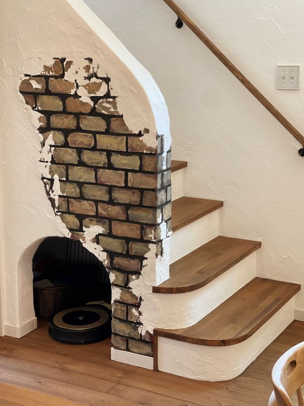 Wooden staircase with a curved white wall revealing exposed brick in an alcove; a robot vacuum sits inside the lower archway.