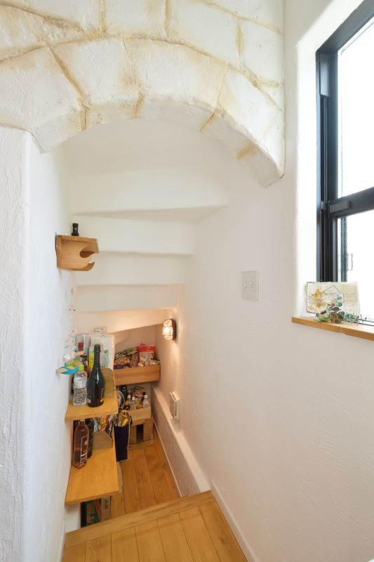Narrow under-stair pantry with wooden shelves filled with bottles, snacks, and small items, light fixture on the wall, white plaster walls, and a window to the right.