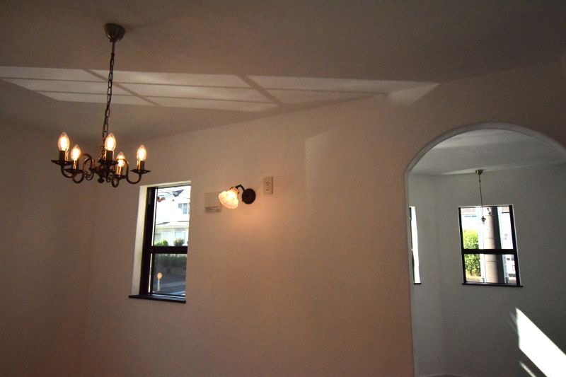 White room with a hanging wrought-iron chandelier, two dark-framed windows, and an arched doorway to a sunlit adjacent space.